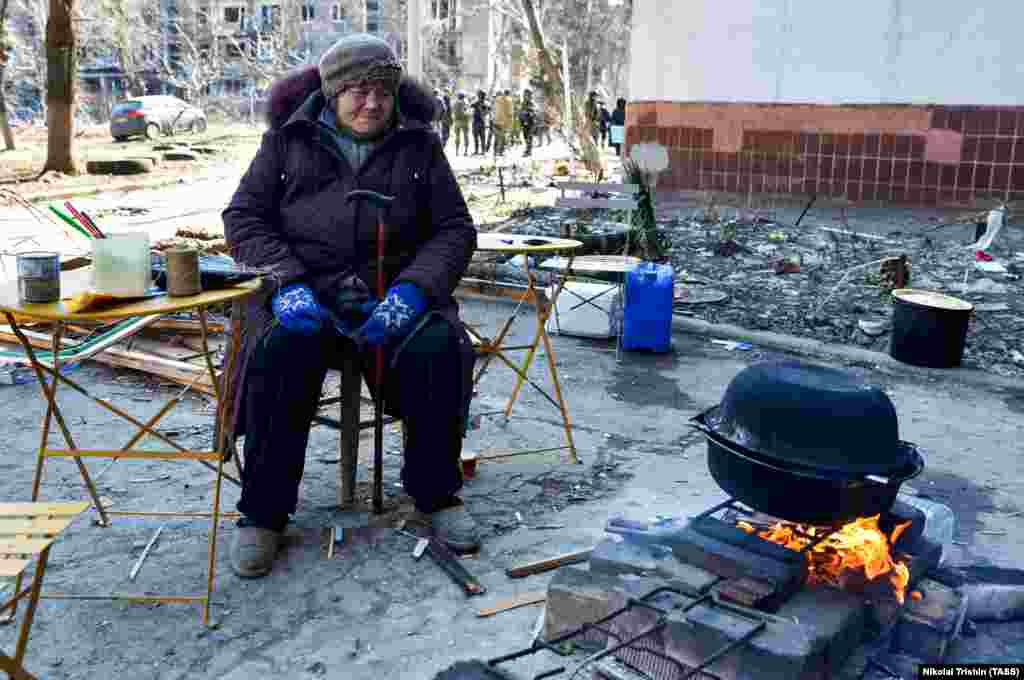 A woman cooks over an open fire near a residential area damaged by Russian shelling. Some must melt snow in order to have water to drink.