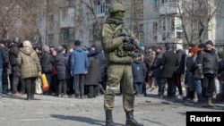 A Russian soldier stands next to local residents waiting in line for humanitarian aid in the besieged southern Ukrainian port of Mariupol. 