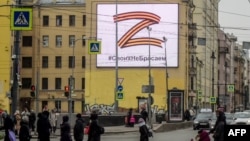 Pedestrians in St. Petersburg cross a street in front of a billboard supporting the Russian Army's unprovoked invasion of Ukraine. Displaying the letter Z in the colors of the ribbon of Saint George, it also contains a slogan saying: "We don't give up on our people." 