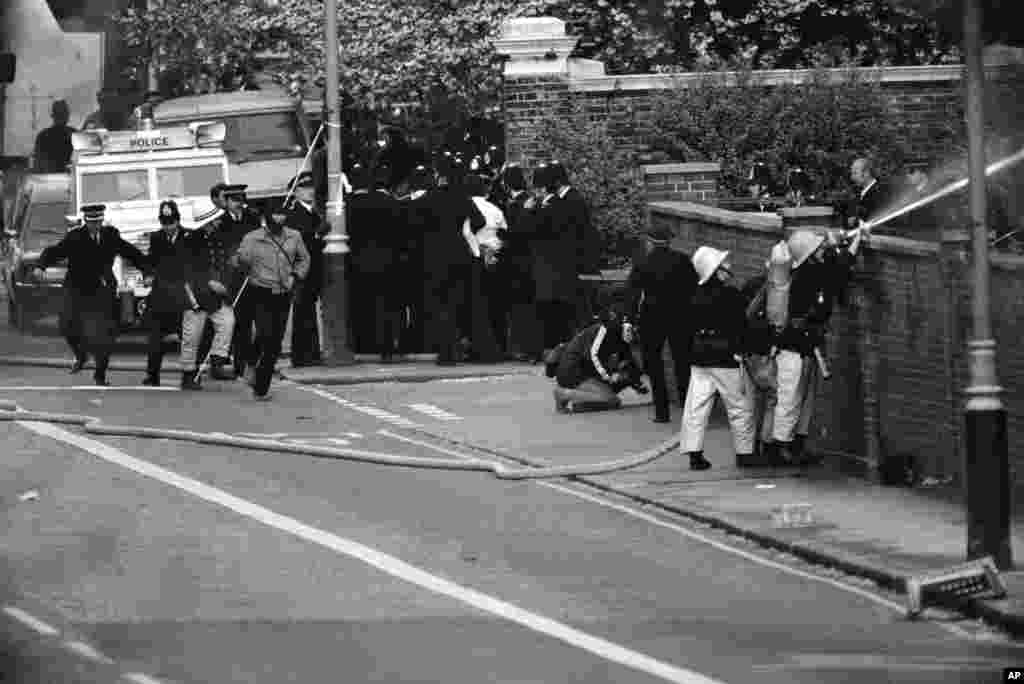 As firefighters aim their hoses at the burning embassy, a group of police officers lead a handcuffed man in a white shirt away. He was thought to have been the sole surviving hostage taker.