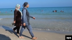 Women walk on a beach wearing burkinis.